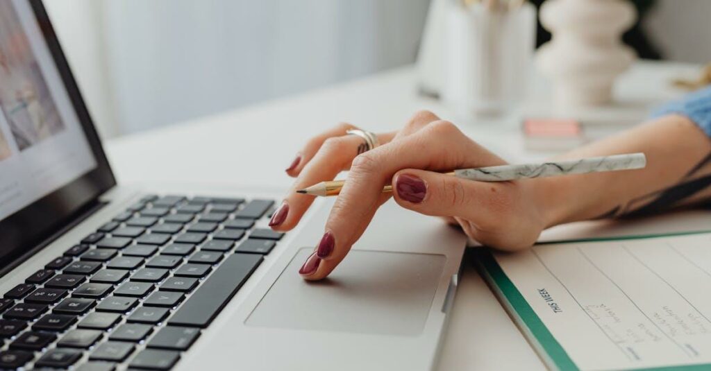 A close-up shot of a woman's hand on a laptop touchpad, holding a pencil, beside a notebook.